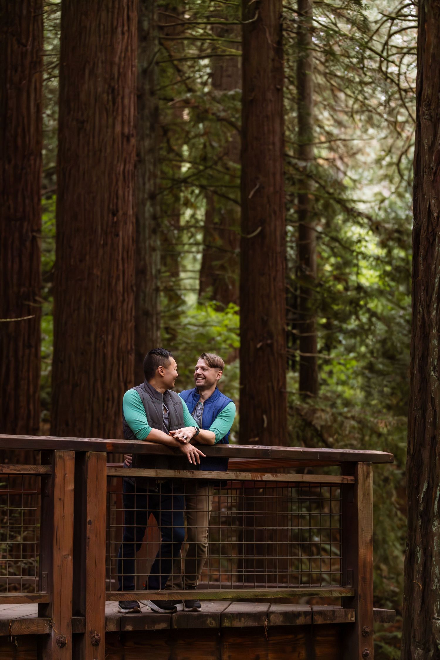 Two people share a romantic moment while sitting on a wooden deck surrounded by towering redwood trees in a forest.