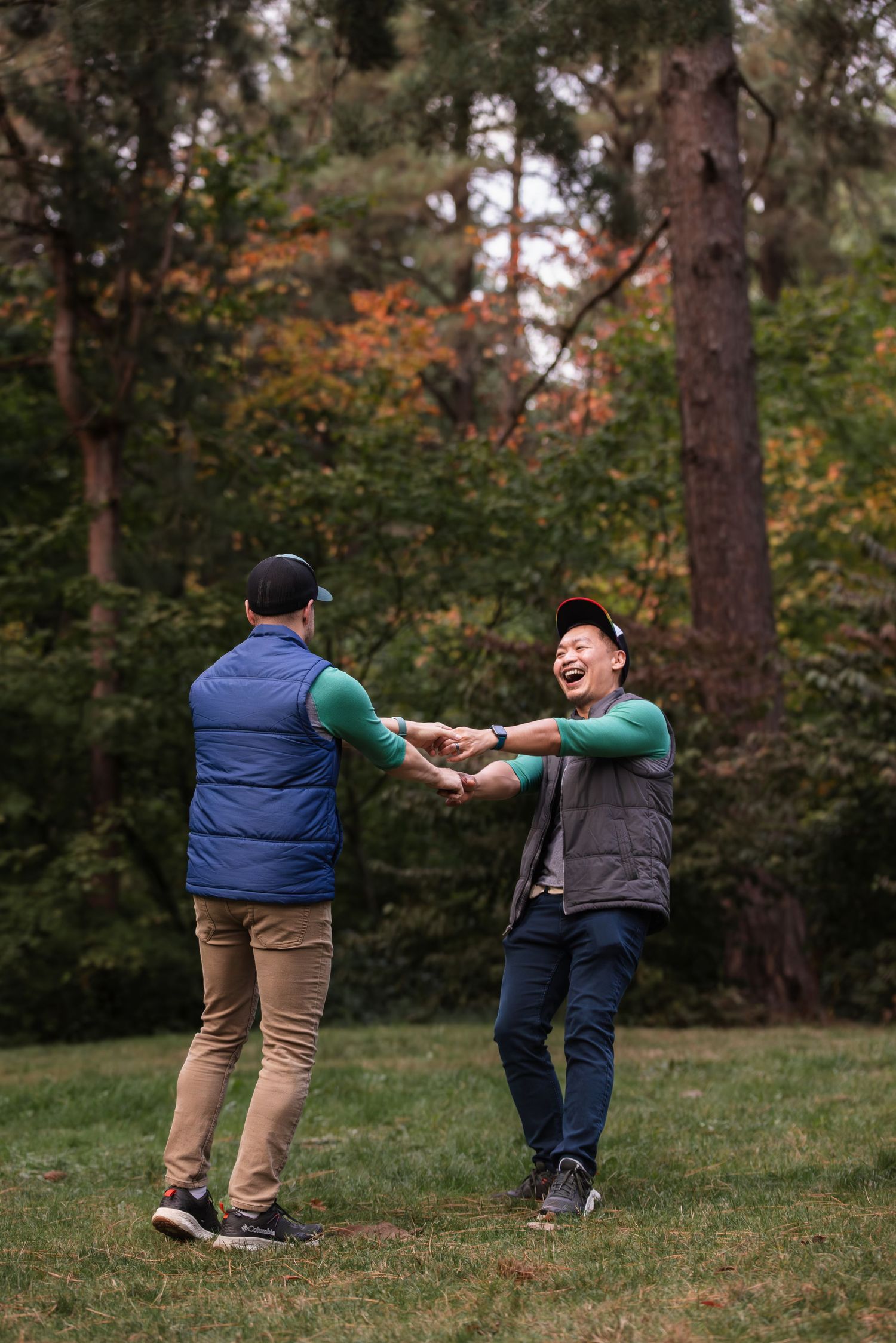 Two people playfully dance together on a grassy hill surrounded by autumn trees in a park setting.