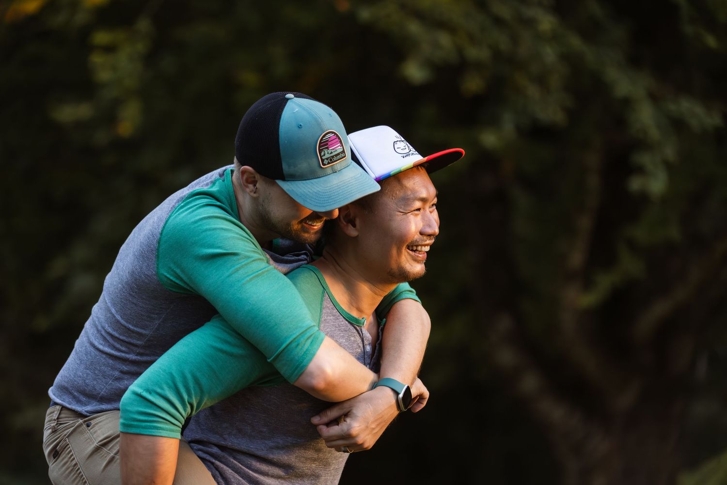 Two people share a joyful piggyback moment outdoors wearing baseball caps and casual attire.