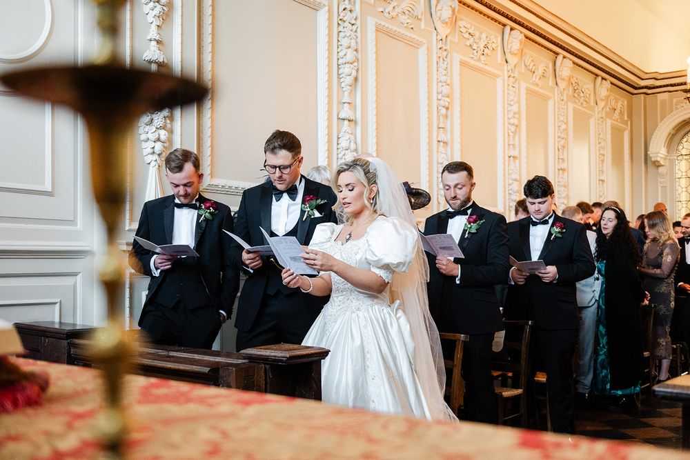 Wedding party members in formal attire stand in an elegant ballroom with ornate walls and chandeliers.