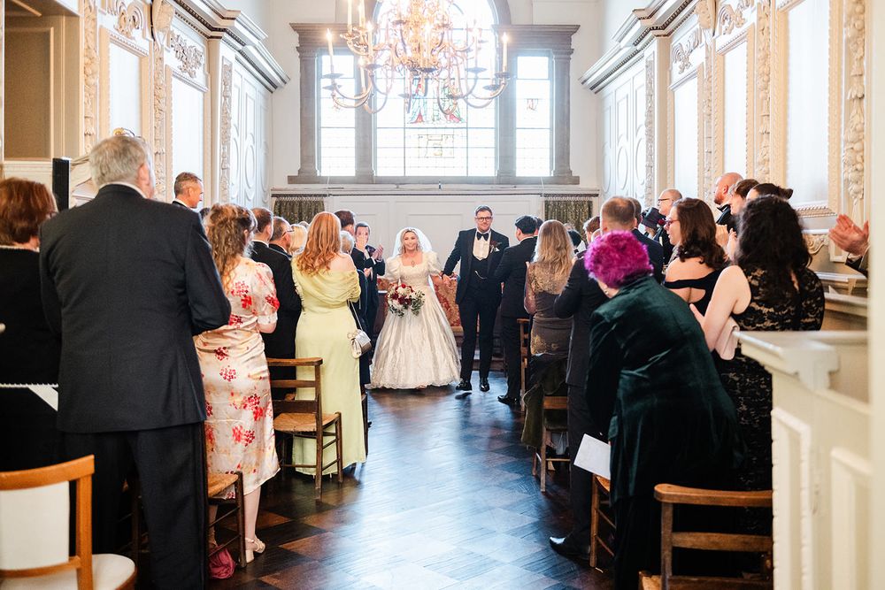 Wedding guests seated at long tables in an elegant white ballroom with crystal chandeliers overhead.