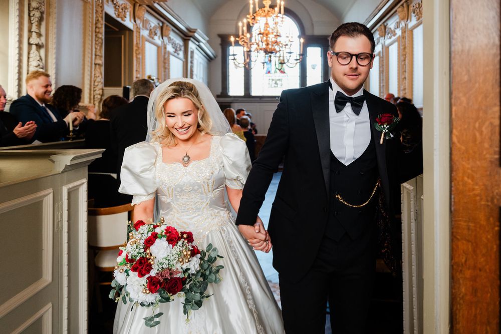 Elegant wedding party walks down an ornate hallway with crystal chandeliers and red floral bouquets.