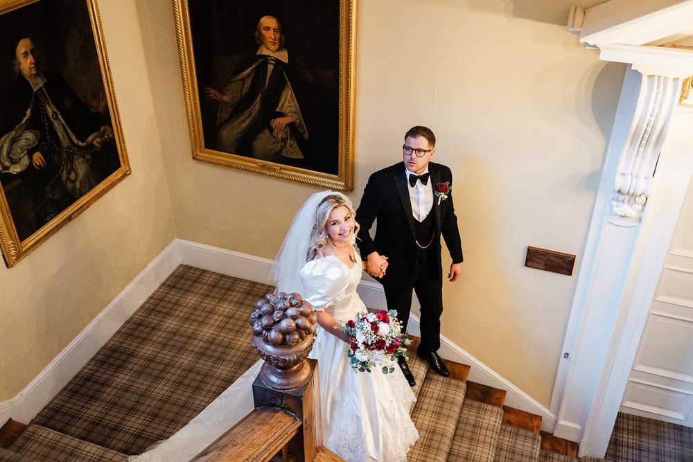 A wedding couple walks down an elegant hallway lined with framed portraits and artwork.