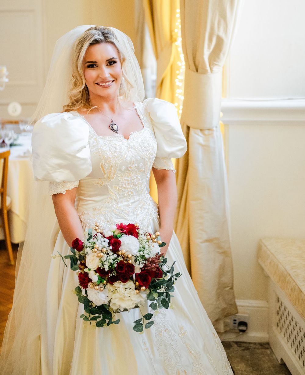 A bride in a white dress with puff sleeves holds a burgundy and white floral bouquet in an elegant room with gold curtains.