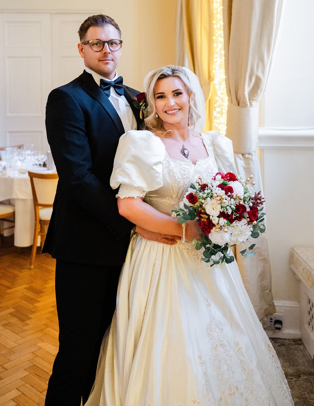 A couple poses together at their wedding, with the bride in a white dress holding a burgundy bouquet against a golden backdrop.