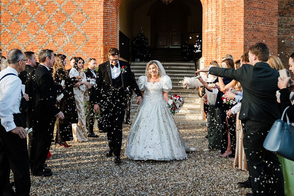 Newlyweds walk through a shower of confetti outside a brick church while wedding guests line the path.
