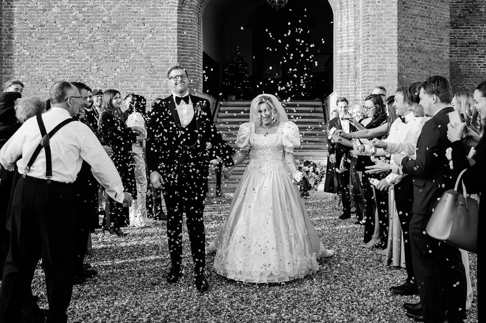 Wedding guests throw confetti over newlyweds outside a brick building during a joyful celebration.