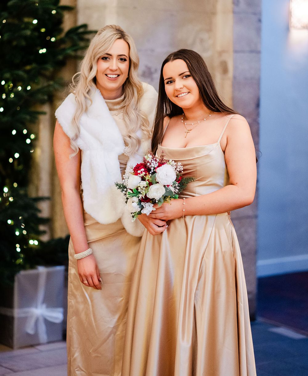 Two people in champagne-colored formal dresses pose together holding a winter floral bouquet near a decorated Christmas tree.