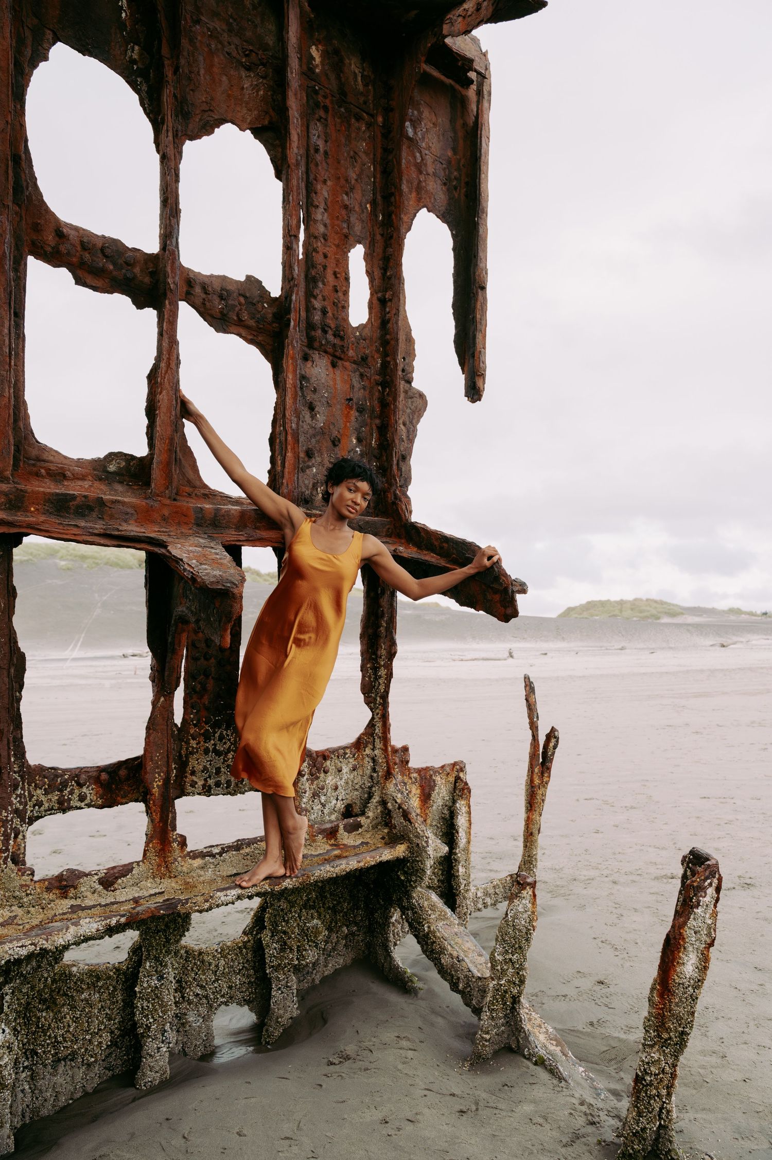 Woman in orange dress posing gracefully on a rusty shipwreck on a sandy beach with overcast skies.
