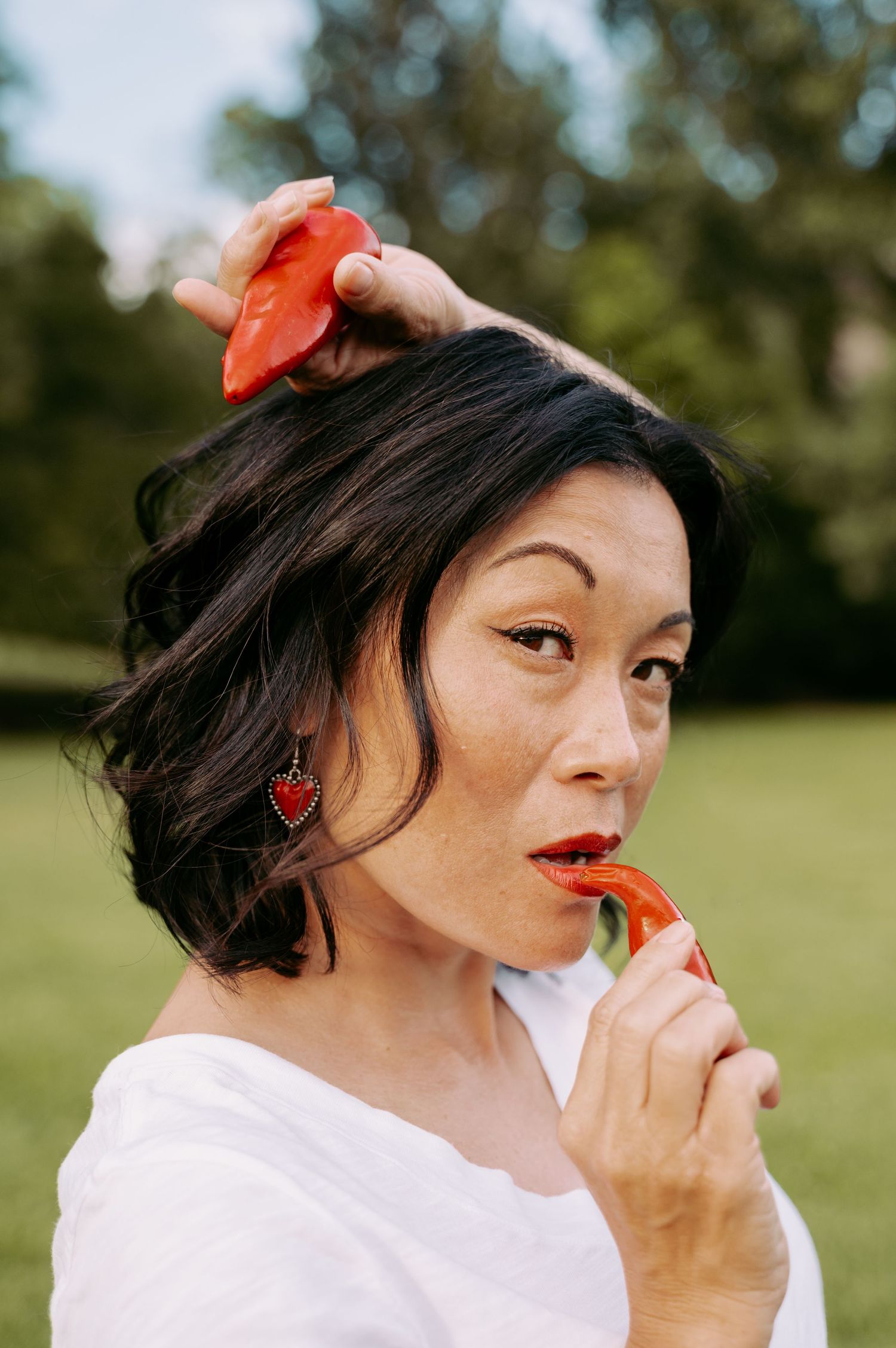A person wearing a white shirt enjoys eating a fresh strawberry in an outdoor setting with green foliage in the background.