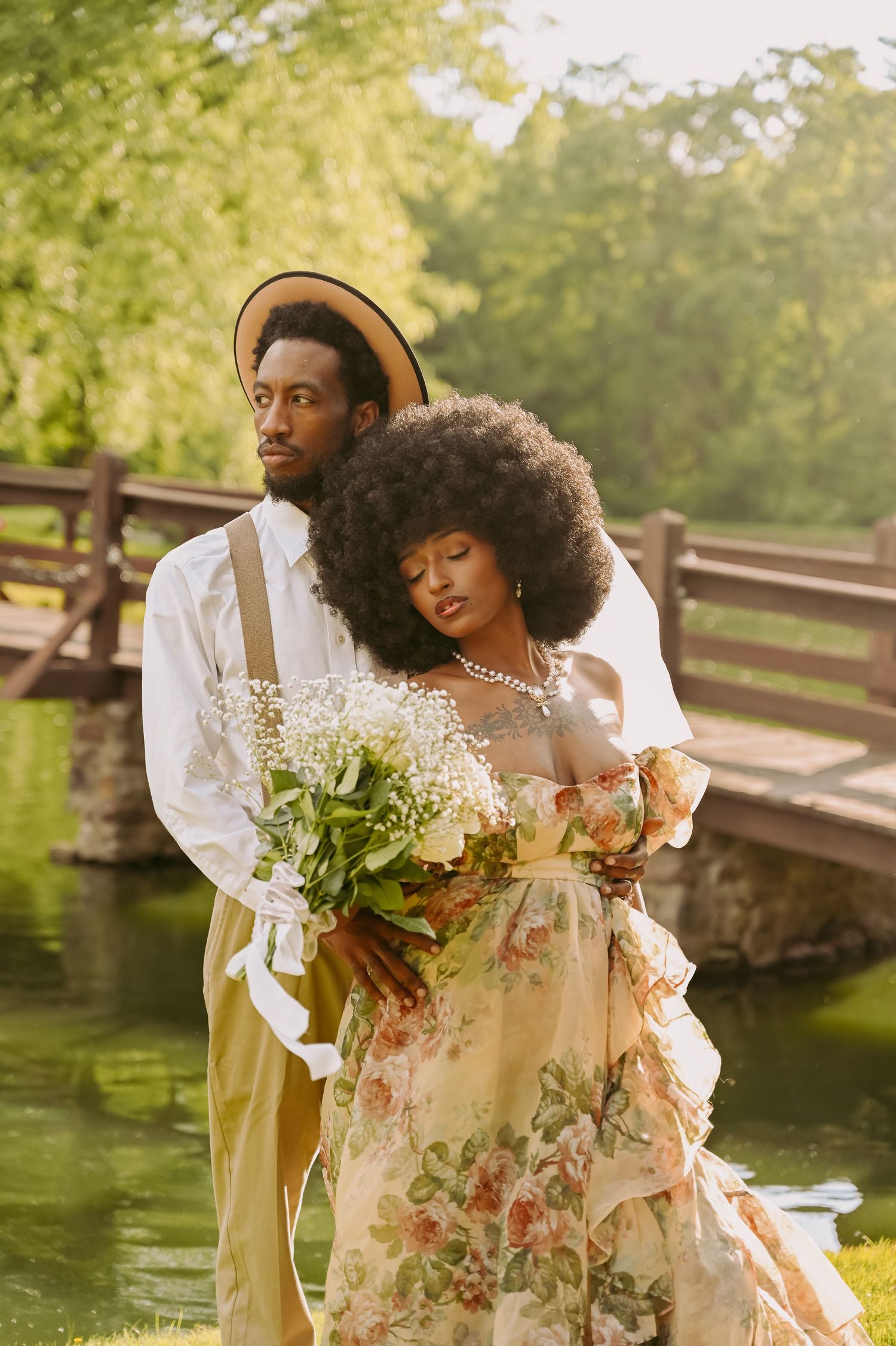 A Black couple poses romantically outdoors near a wooden bridge, she wears a floral gown holding a bouquet, he wears a straw hat.