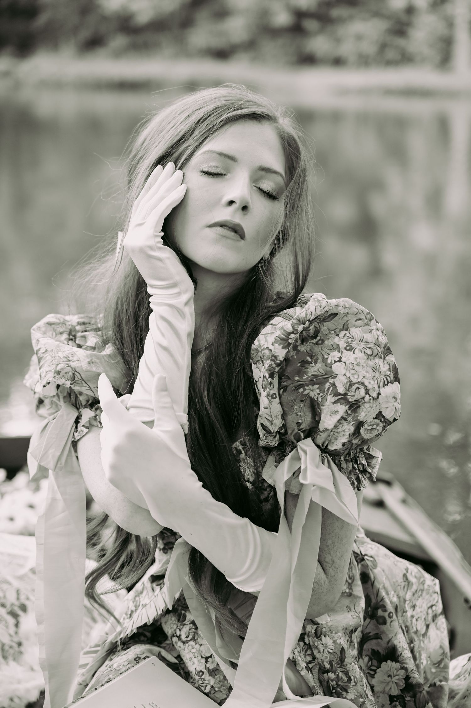 Black and white portrait of a woman in a floral Victorian dress and long gloves, eyes closed, posed elegantly outdoors.