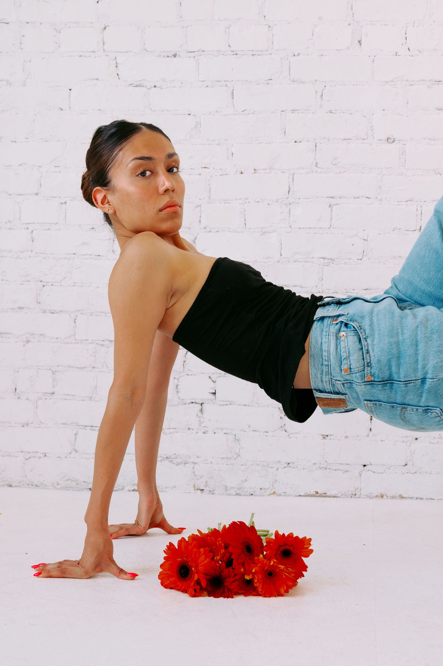 Woman in black tube top and jeans poses near red gerbera daisies against white brick wall.