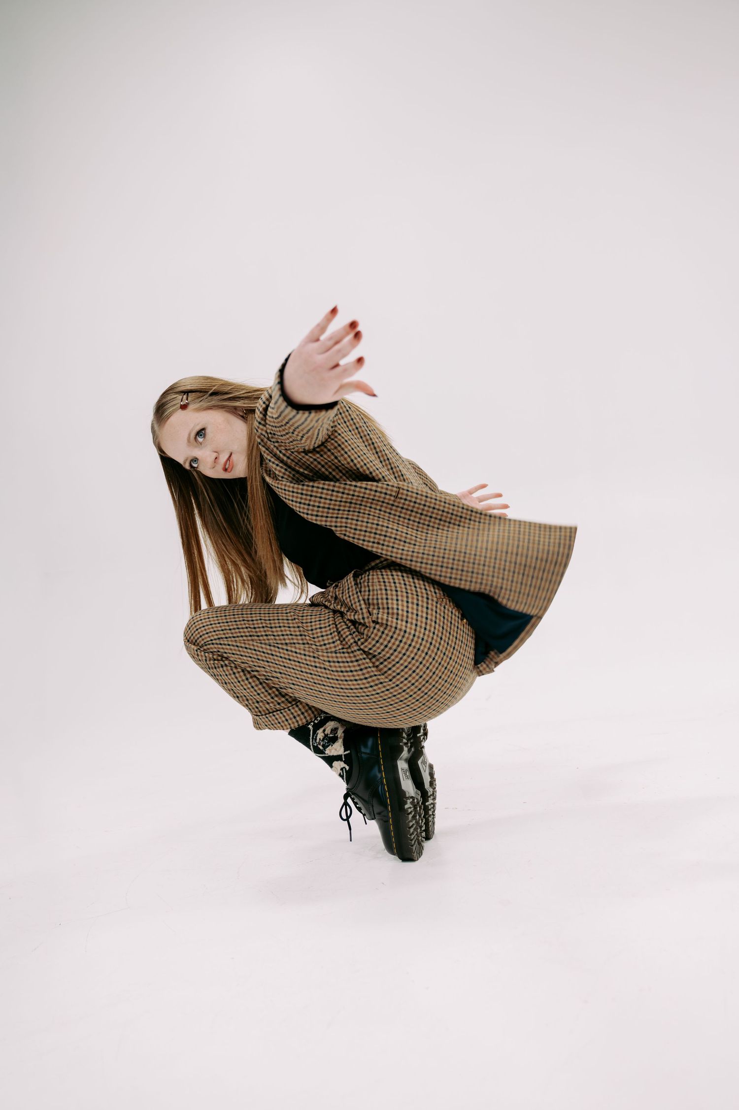 Young woman in plaid oversized coat and black boots crouching dynamically against white background.