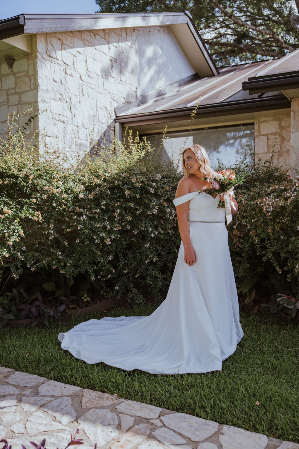 Elegant white wedding dress with flowing train photographed in outdoor garden setting with stone wall backdrop.
