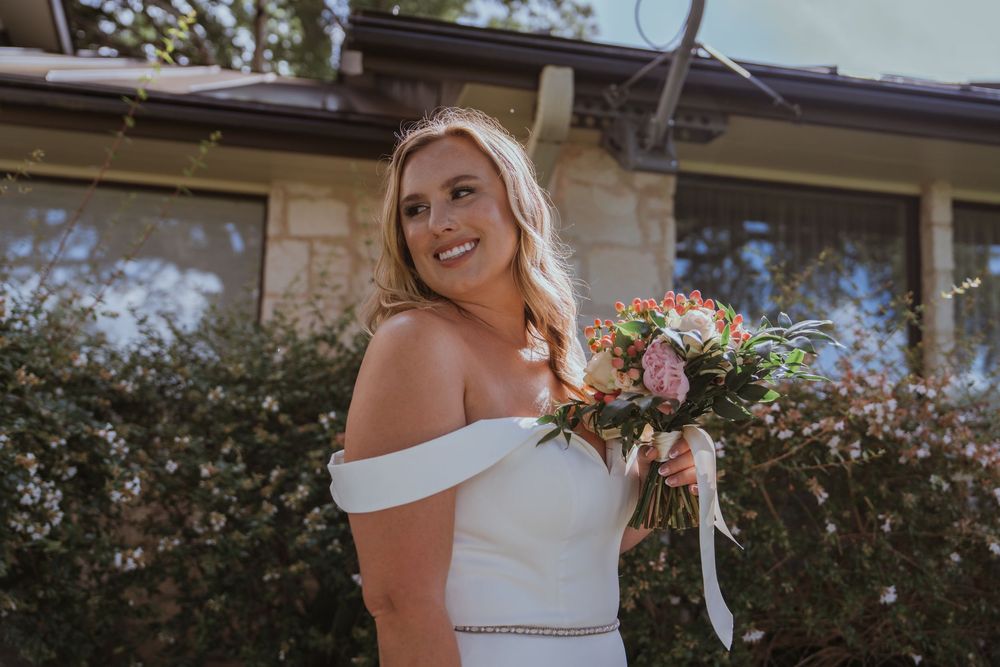 Bride in off-shoulder white gown holding colorful bouquet against stone building exterior.
