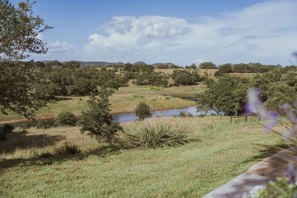 Rolling green hills and blue sky landscape with scattered trees and pond in distance.