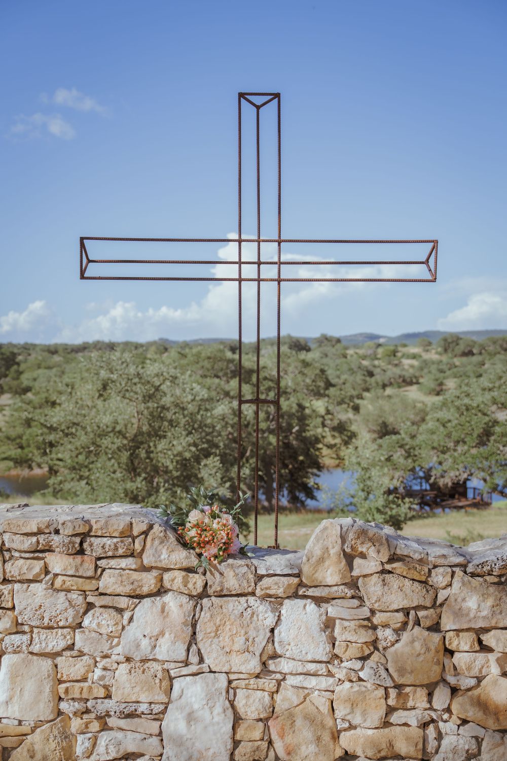A minimalist metal cross stands atop a rustic stone wall against a bright blue sky.