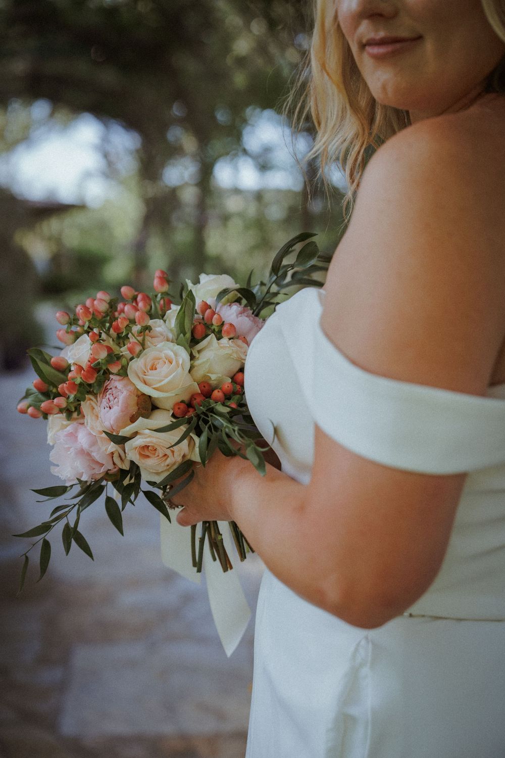 A delicate bridal bouquet features peach roses and greenery against a white dress.