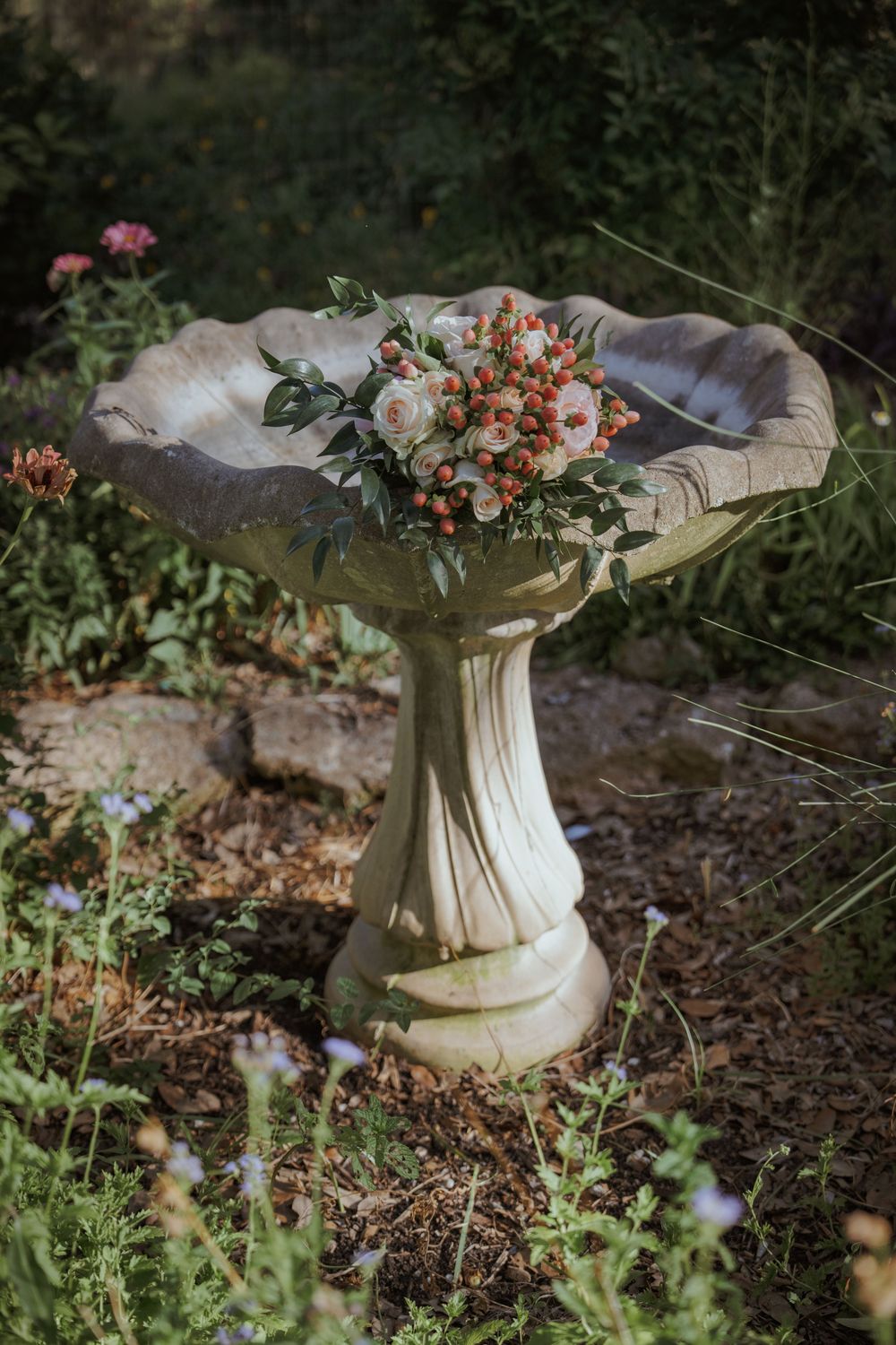 A decorative stone birdbath adorned with floral arrangements sits in a garden setting.
