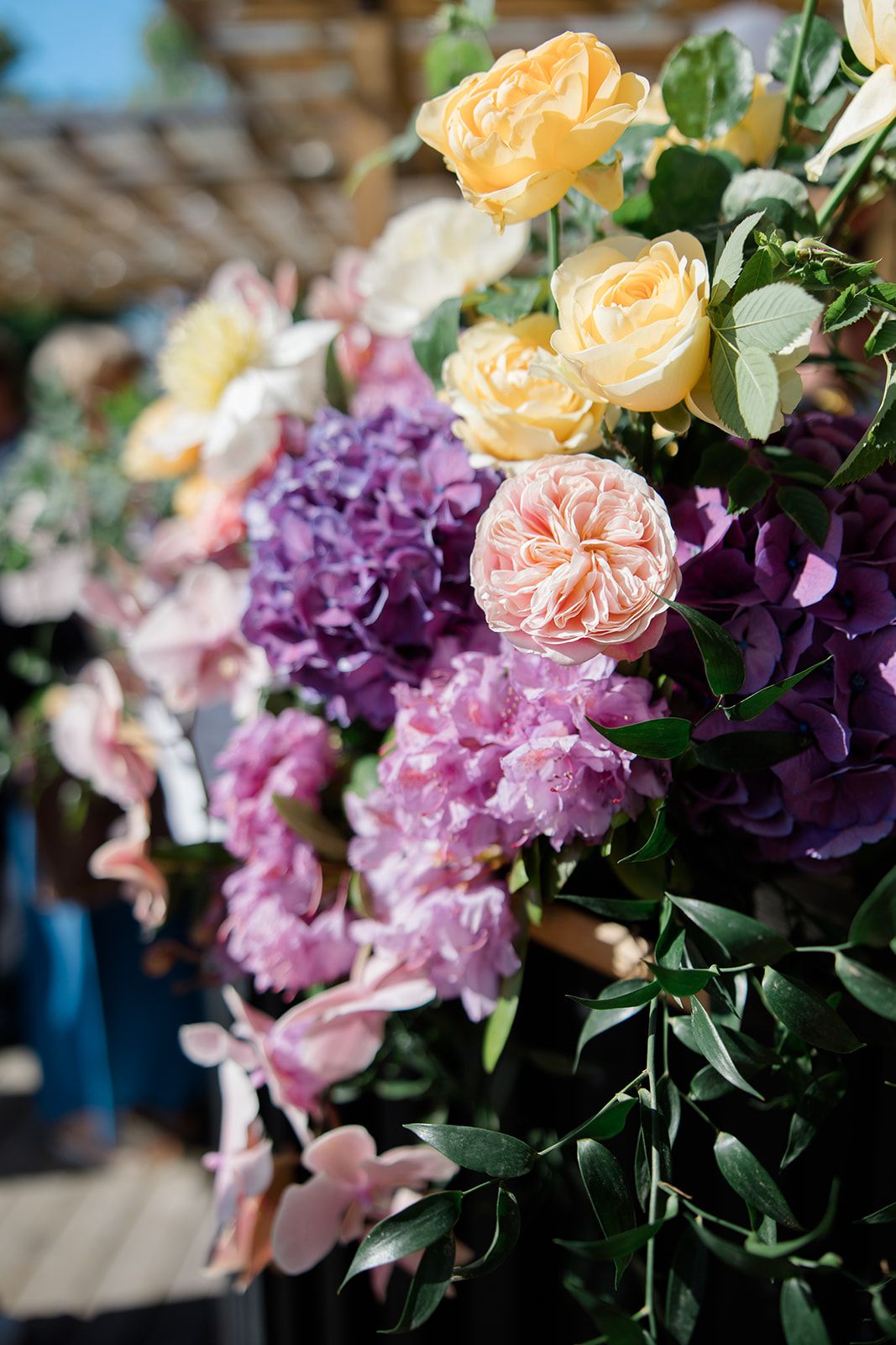 Beautiful floral arrangement featuring yellow roses, pink garden roses, and purple hydrangeas with lush greenery.