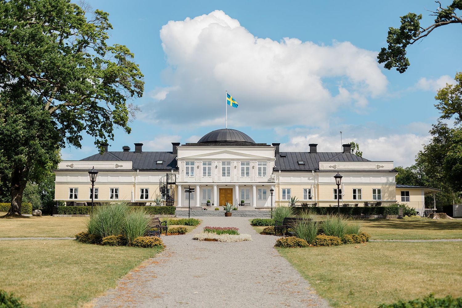 A panoramic view of a grand white neoclassical mansion with manicured gardens and lawns under a partly cloudy sky.