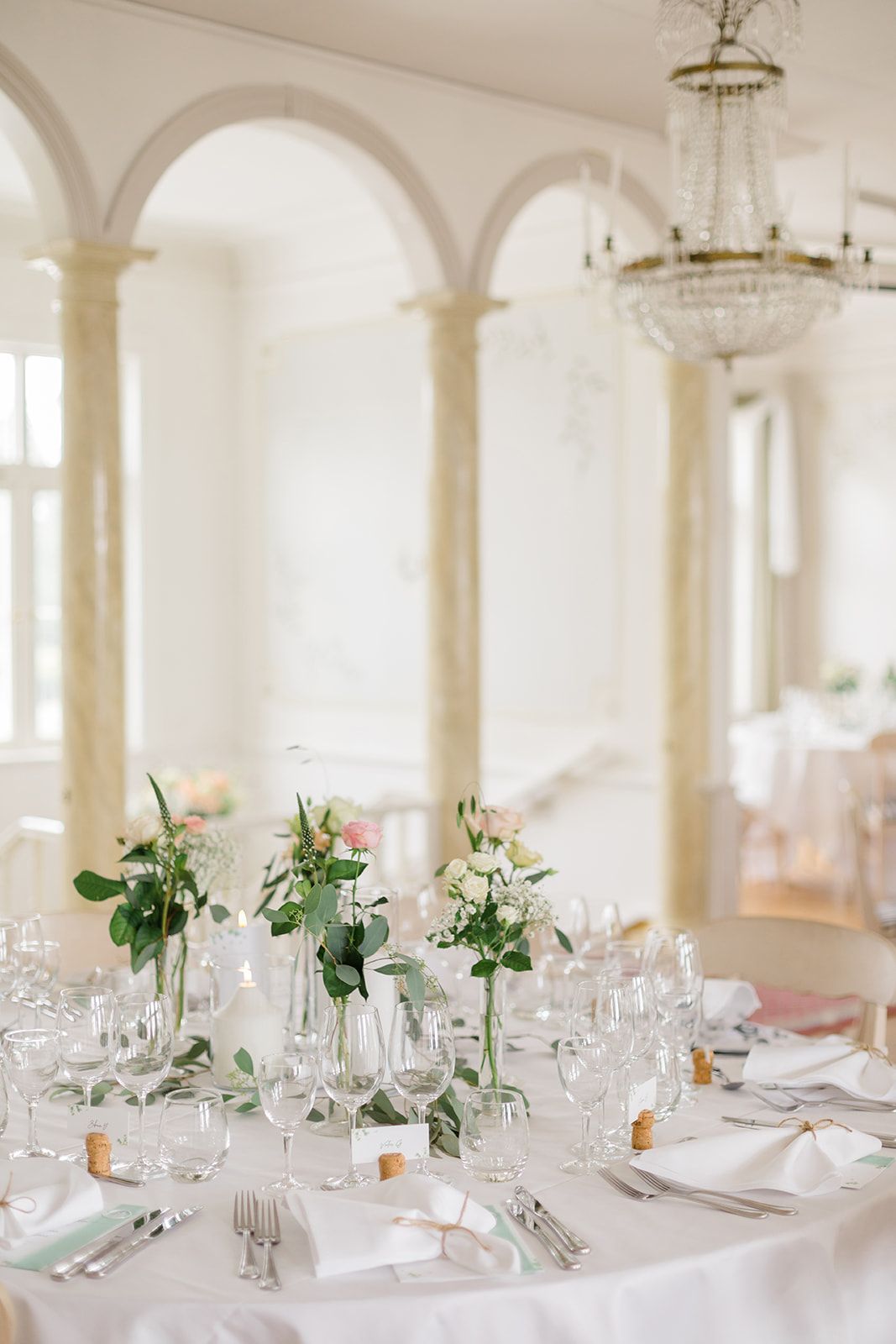 Elegant white wedding reception tables set with crystal glassware and delicate green floral centerpieces in a bright room.