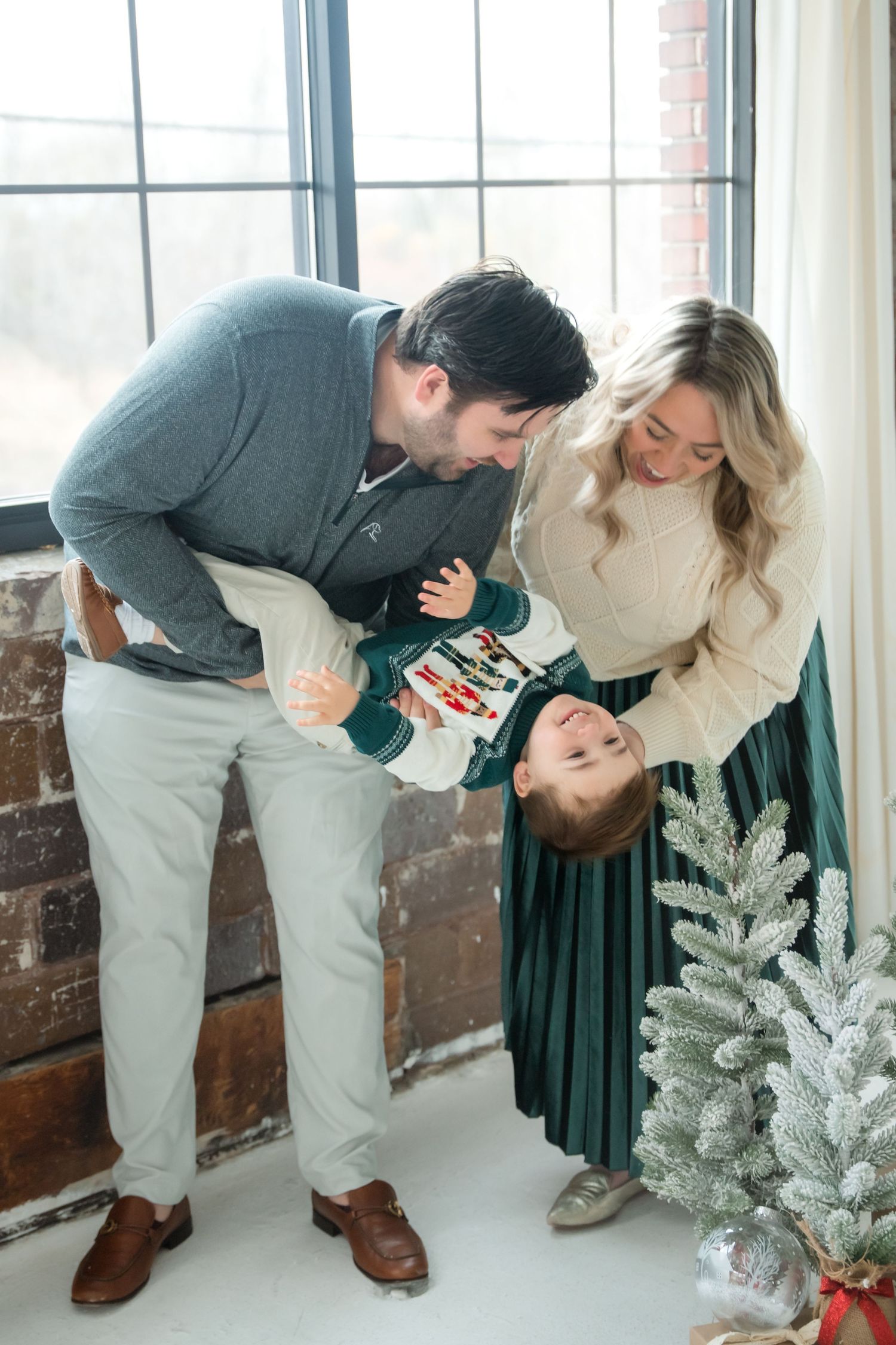 A family shares a joyful moment together near a white flocked Christmas tree by a brick wall and window.