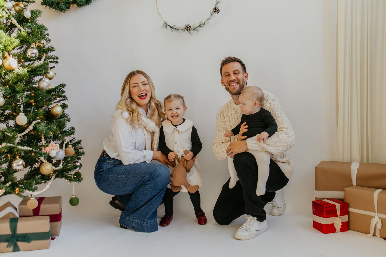 A family poses together by a decorated Christmas tree with wrapped presents on the floor during a holiday photoshoot.