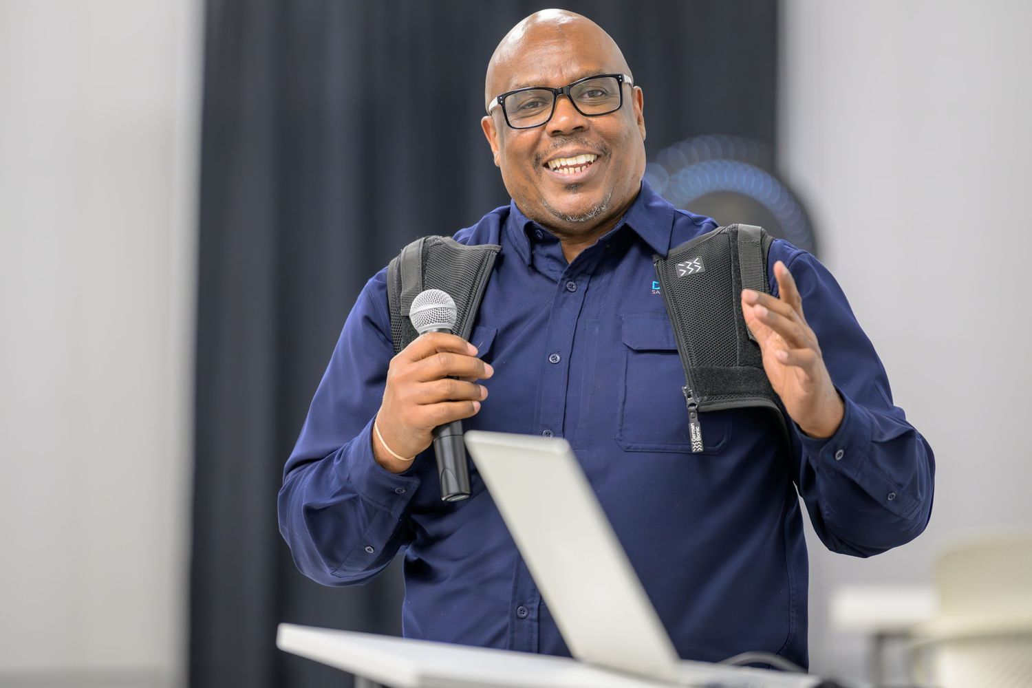 A person in a navy blue jacket gestures while speaking into a microphone during a presentation.