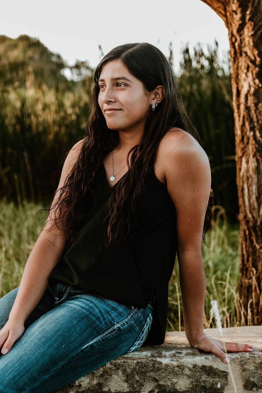 Person in dark top and jeans poses on rocky surface surrounded by pine trees.