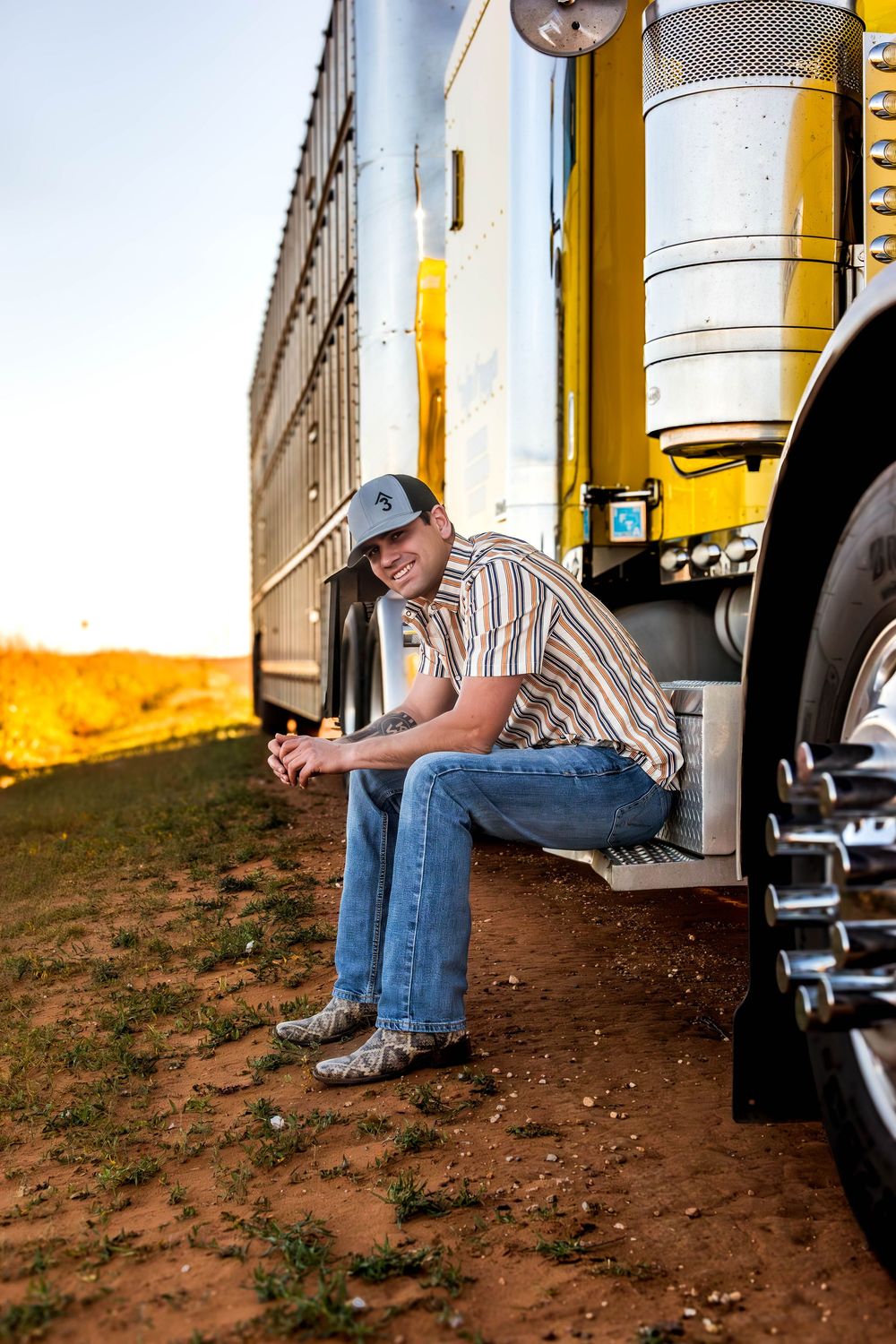 Person in striped shirt and jeans sits on steps of a yellow semi truck.