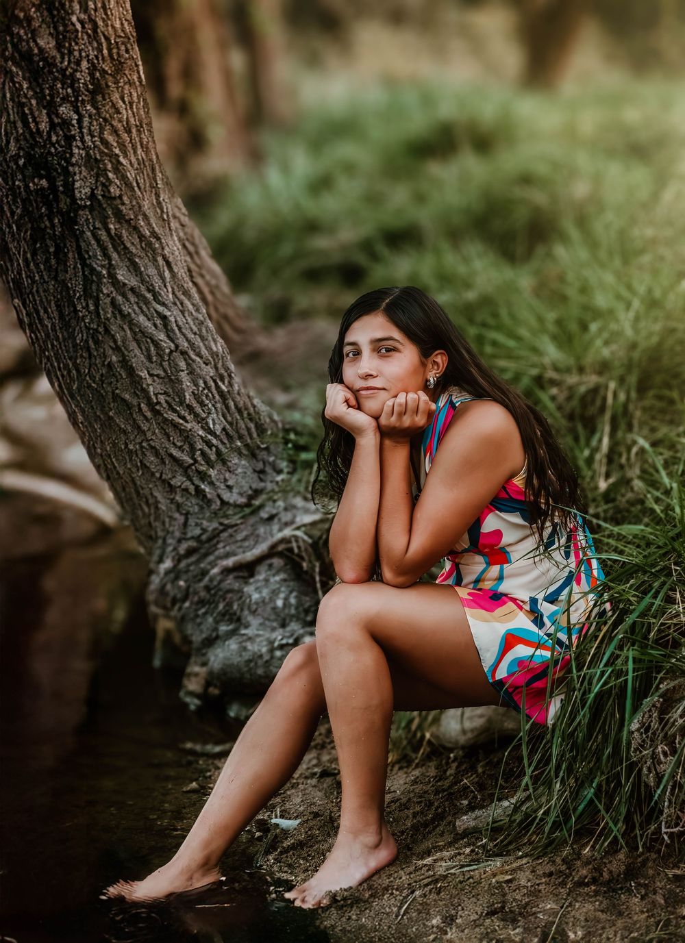 Person in floral dress sits barefoot near tree trunk in natural outdoor setting.