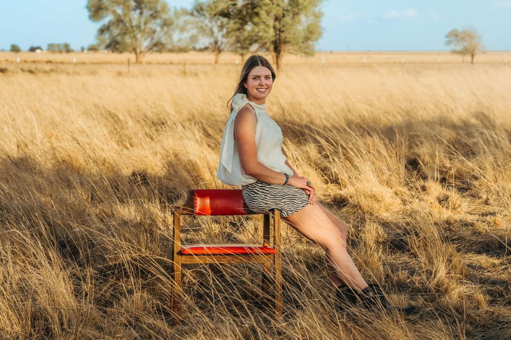Woman sitting on a red chair in golden grassland with trees in background during sunset.