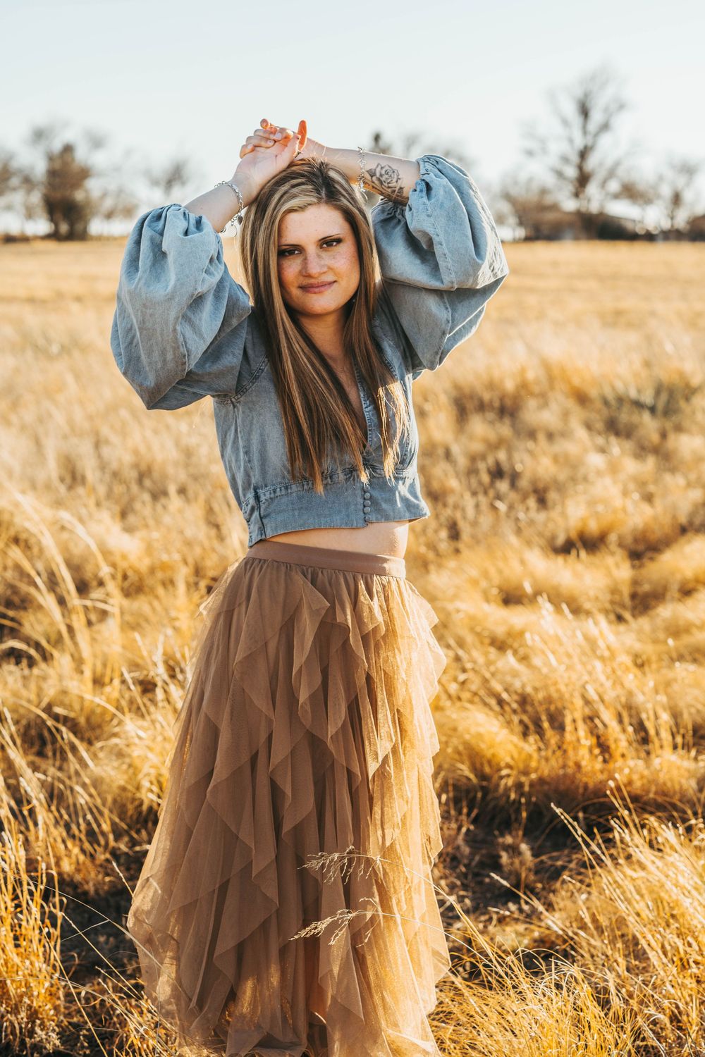 A denim jacket and flowing brown tulle skirt create a stylish contrast against golden autumn grass at sunset.