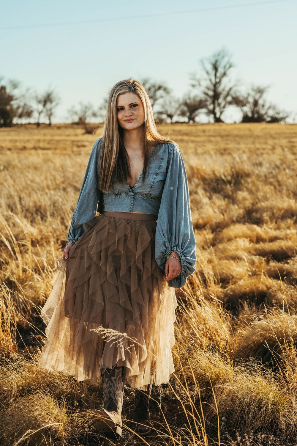 Person in denim top and brown tulle skirt poses in golden field at sunset during outdoor photoshoot.
