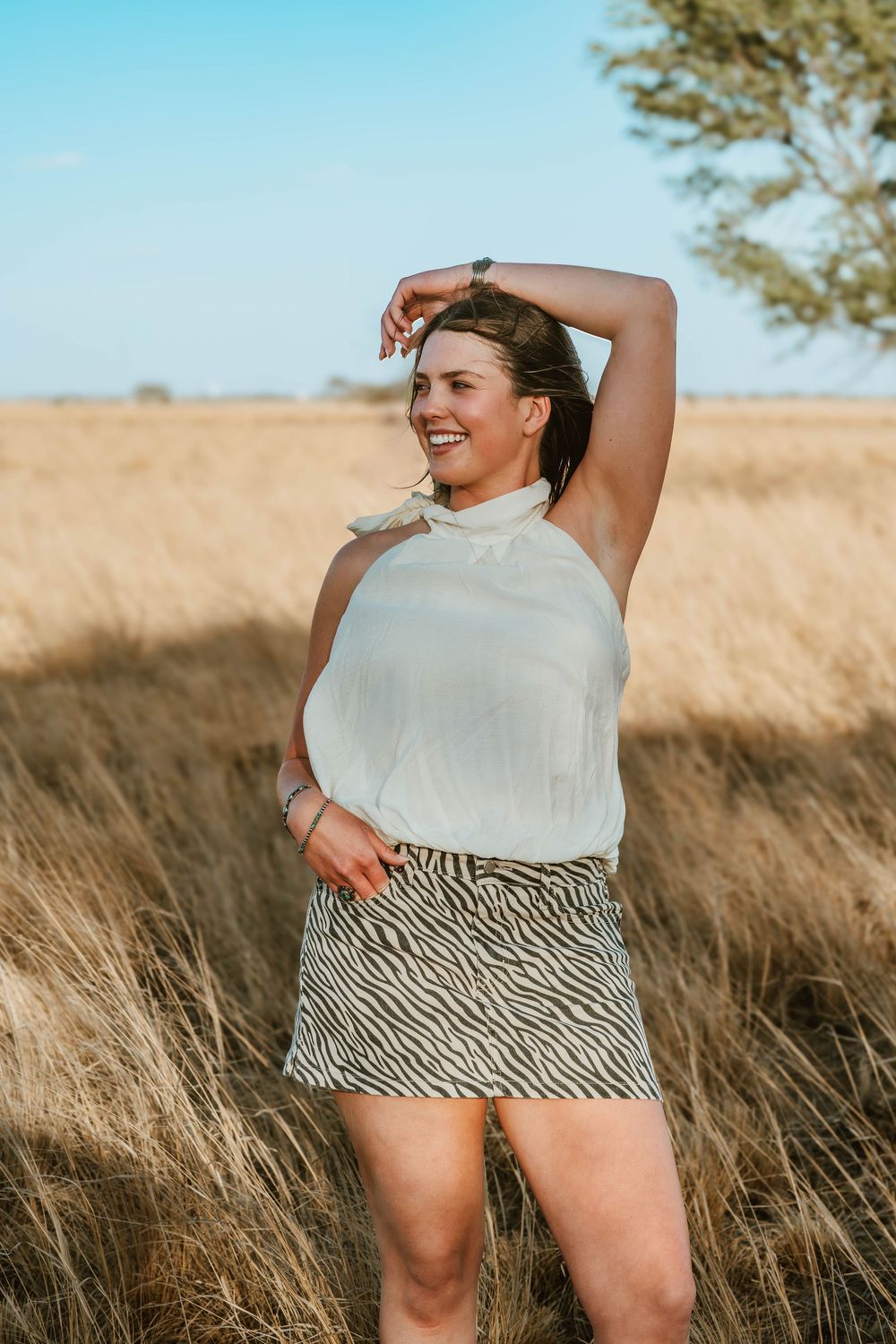 Woman in white halter top and zebra print mini skirt smiling in golden grass field under blue sky.