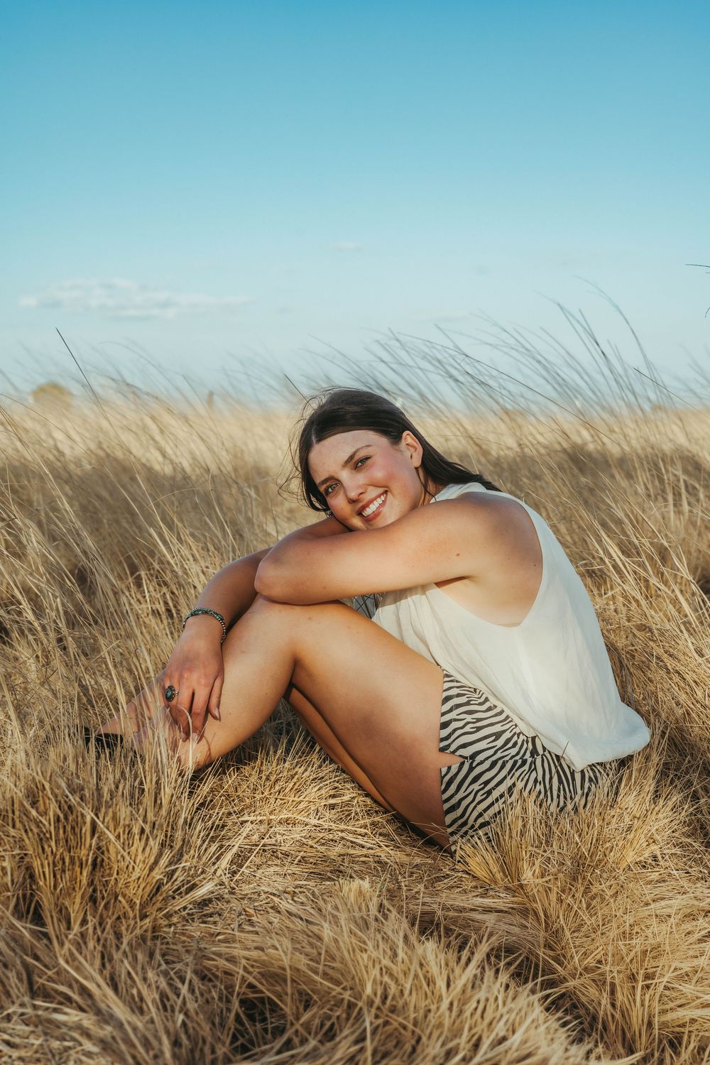 Smiling young woman in white top and striped shorts sitting relaxed in tall golden grass field.