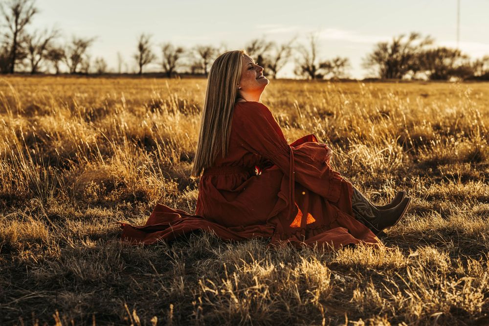 Person in brown coat sits in golden grass field at sunset while silhouetted trees line the horizon.