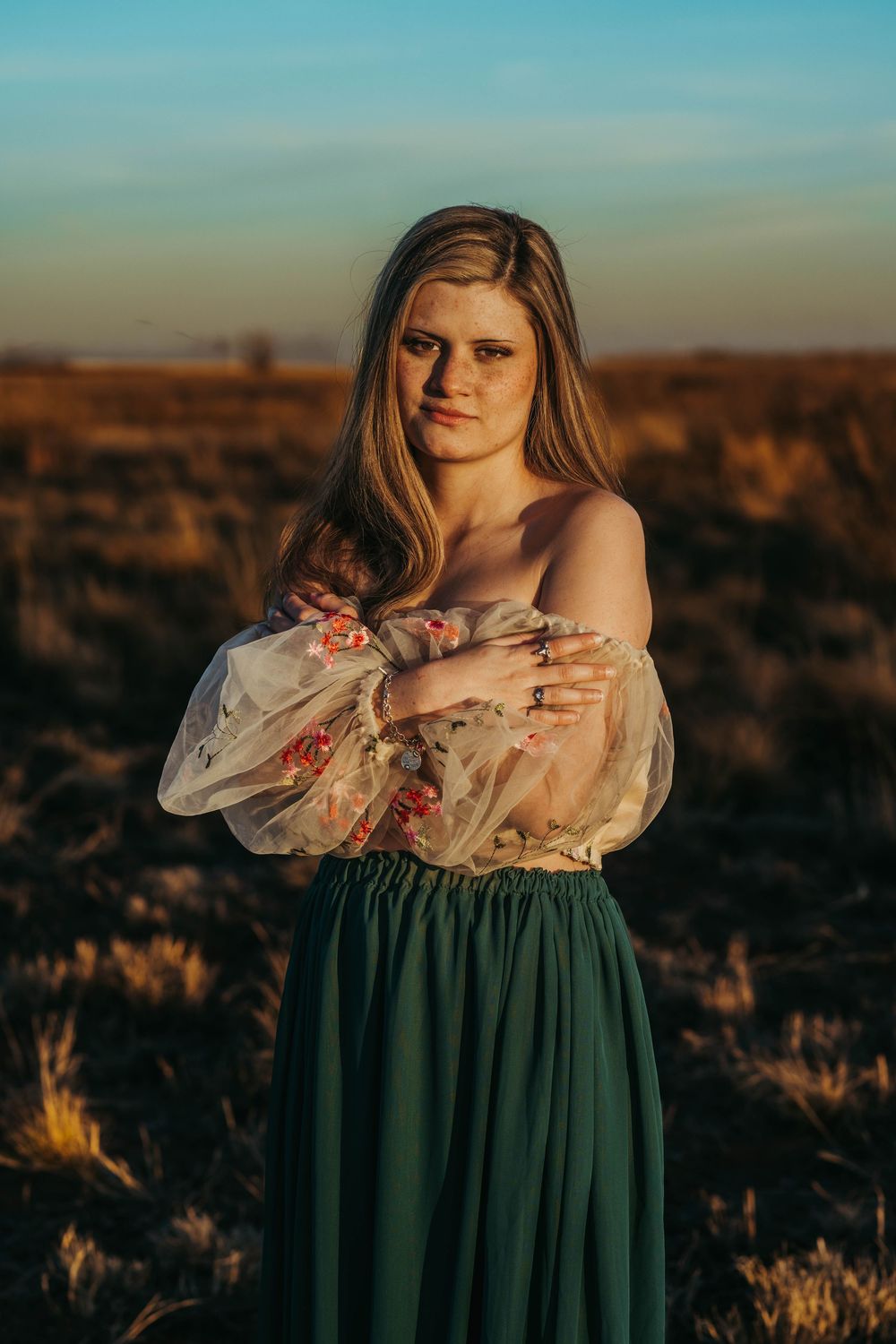 A person in a floral off-shoulder top and dark green skirt poses in a field at golden hour.