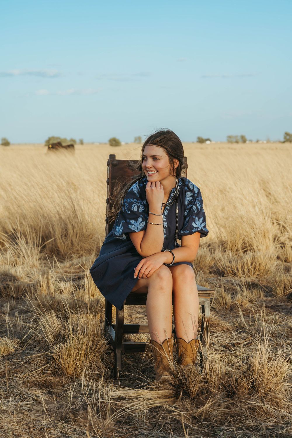 Young woman in blue floral dress and cowboy boots sitting on wooden chair in golden wheat field.
