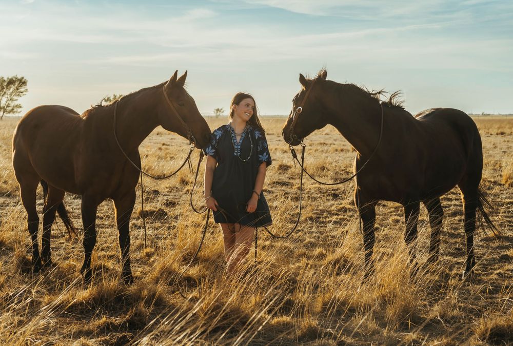 Young woman standing between two brown horses in a golden field at sunset.