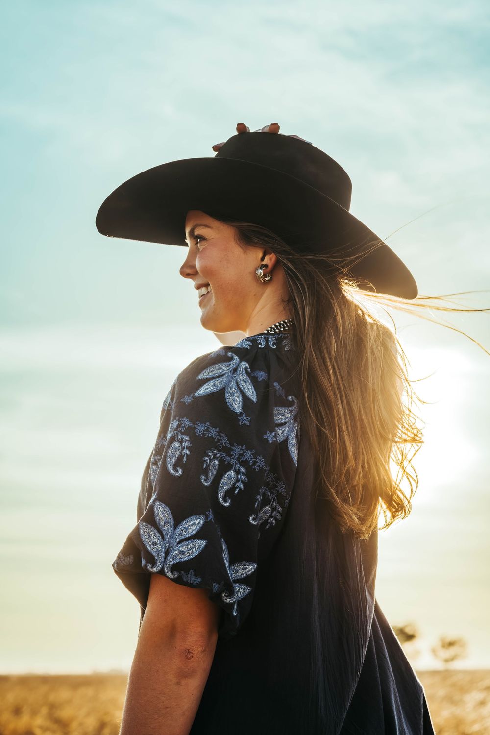 Smiling woman in black wide-brim hat and floral blouse stands in golden field at sunset, hair blowing in wind.