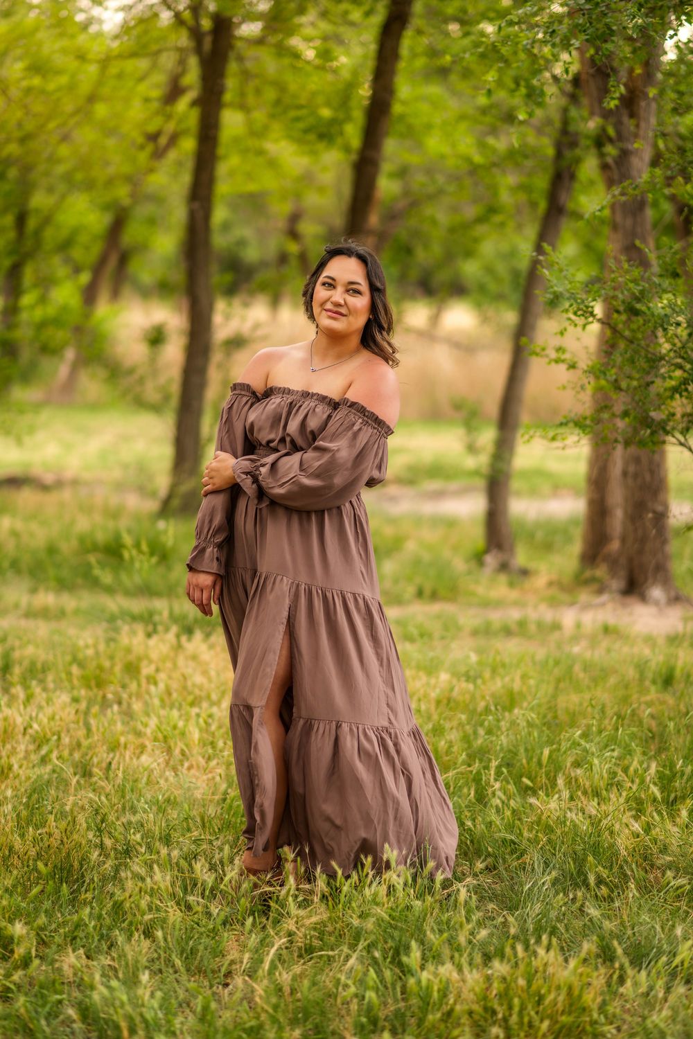Woman in brown off-shoulder maxi dress smiling in a lush green park surrounded by tall trees.