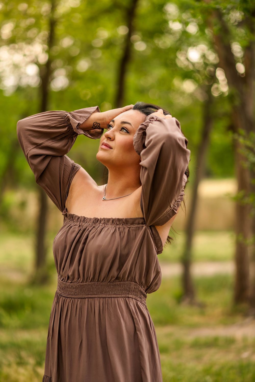 Woman in brown off-shoulder dress looking up in forest.