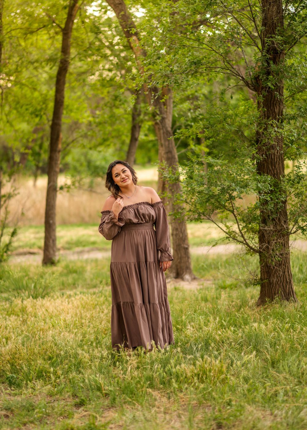 Woman in brown off-shoulder maxi dress smiling in lush green forest meadow during golden hour outdoor portrait.