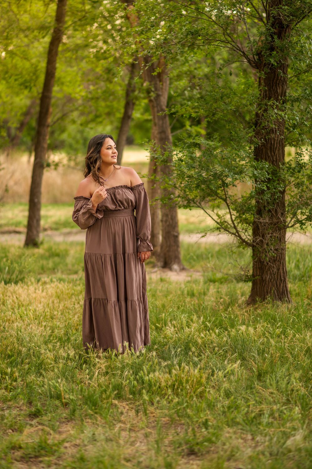 Woman in brown off-shoulder maxi dress standing among green trees in a serene outdoor setting.