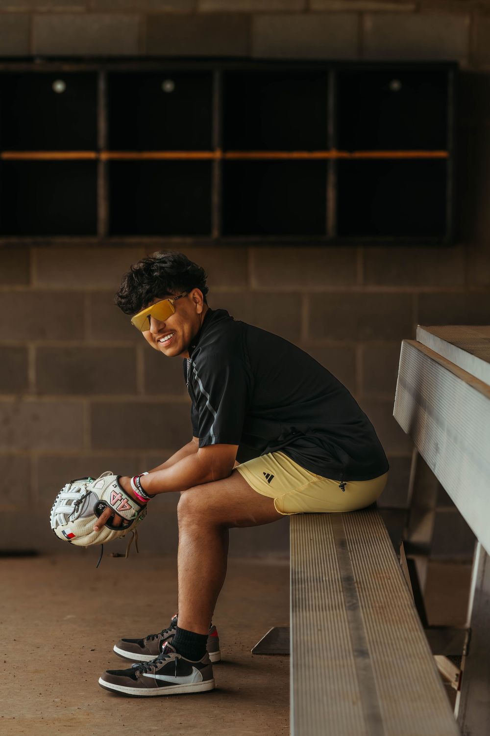 Smiling young baseball player sitting in dugout wearing yellow shorts and sunglasses, holding a fielding glove.