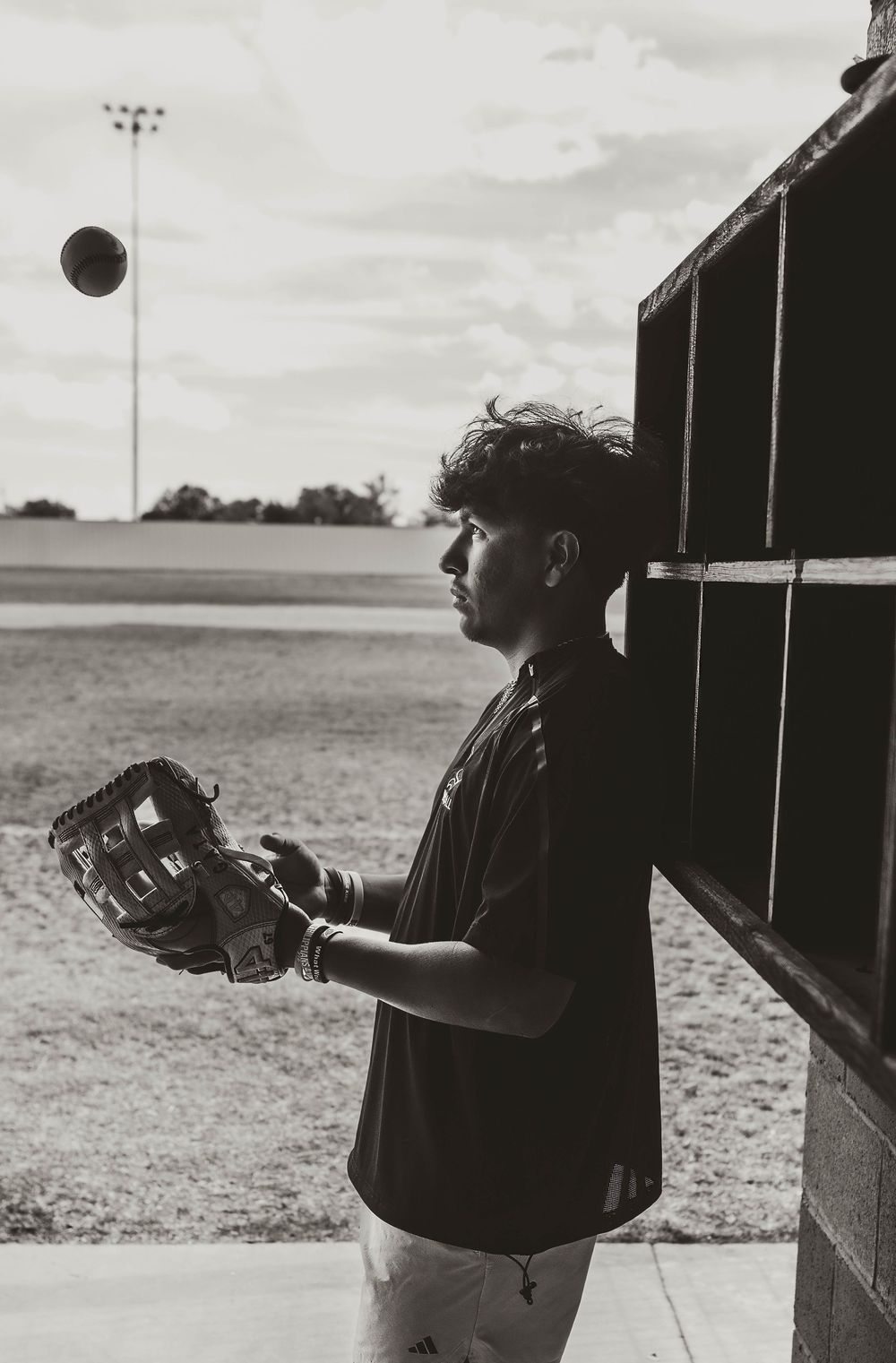 Black and white photo of a young baseball player wearing a mitt, watching a ball in the air near a dugout.