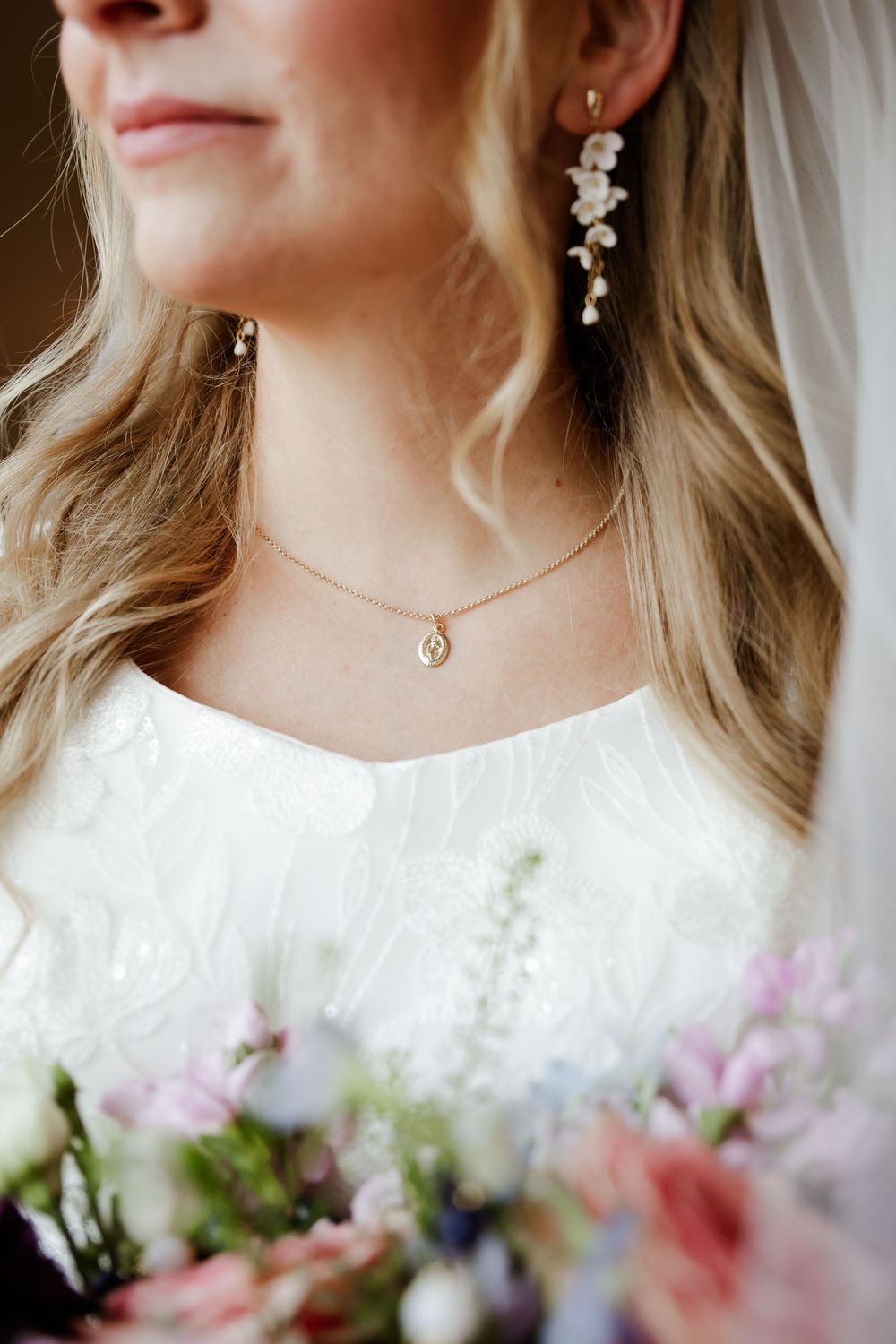 Bride wearing floral drop earrings, gold pendant necklace, and white lace dress holding a pastel bouquet.